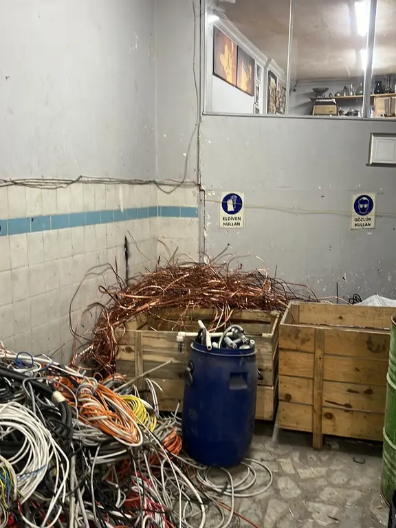 Hızlı Hurda deposunda ahşap kasa içinde toplanmış bakır tel ve ayrı yığılmış tesisat kabloları; duvarda eldiven ve gözlük kullanımı uyarı levhaları